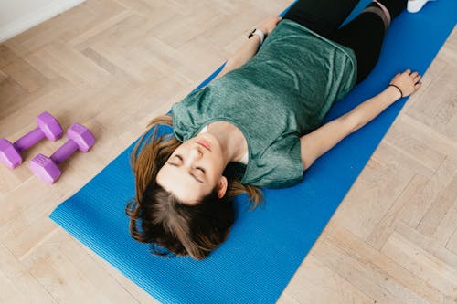 From above of young slender lady in casual clothes and wristwatch resting with closed eyes on blue yoga mat near dumbbells on floor in flat