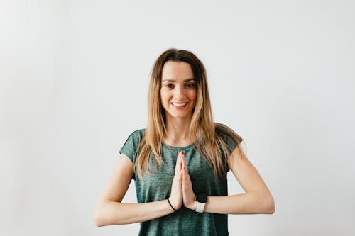 Glad blond woman in casual clothes and smart watch standing in Namaste pose while practicing yoga on white background and looking at camera