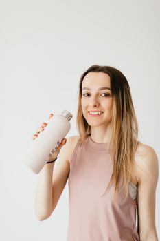 一位穿着休闲装、笑容灿烂的金发女子站在白墙边，手拿一瓶清凉饮料，看着镜头