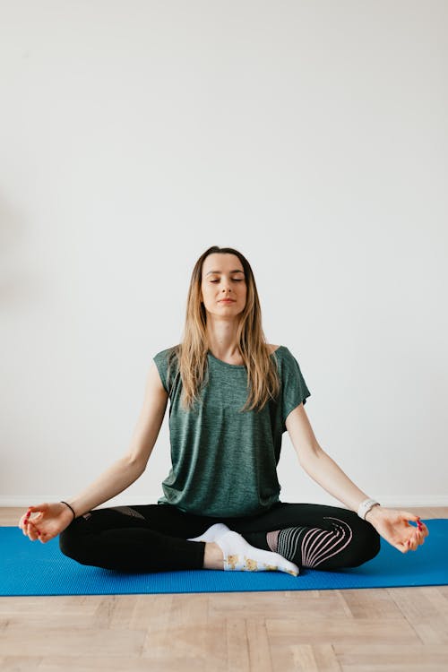Free Calm slim female in sports clothes sitting with crossed legs in Padmasana pose on yoga mat while meditating with closed eyes in flat Stock Photo
