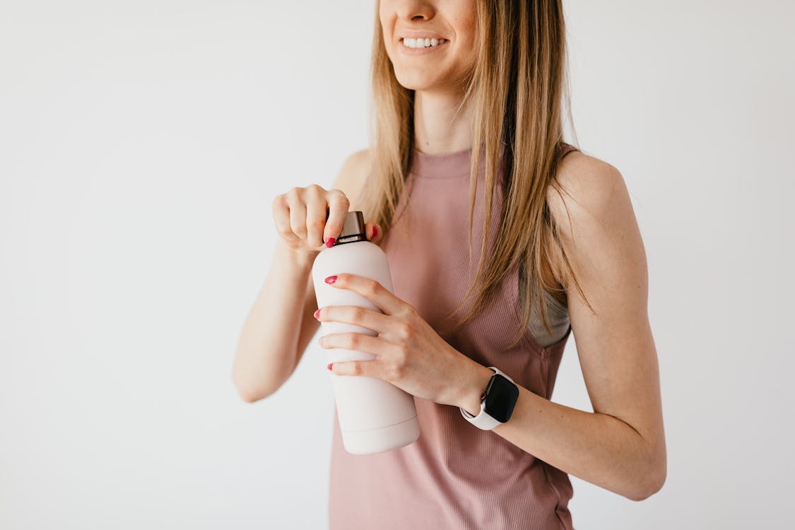Crop faceless young female in casual outfit wearing smart watch opening cosmetic bottle while standing against white background