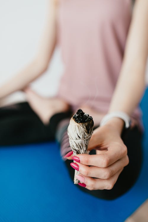 Crop unrecognizable female in casual clothes sitting on blue mat and meditating in lotus position while holding burning natural white sage incense in hand