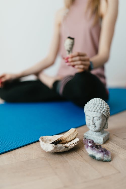 Selective focus of crop faceless young female in casual wear meditating in lotus pose with white sage incense near Buddha head sculpture amethyst crystal and palo santo wood in stone bowl