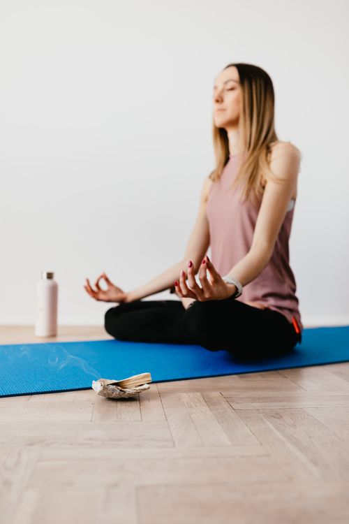 Free Young slim female sitting with closed eyes in Padmasana on yoga mat with water bottle on one side and Bursera Graveolens giving off aroma on other Stock Photo