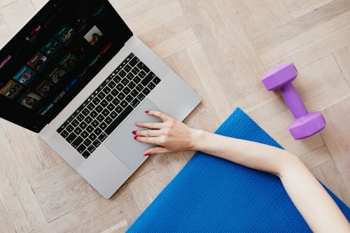 Top view of crop anonymous female looking for video workout courses on laptop while sitting on blue yoga mat with purple dumbbell beside on parquet floor