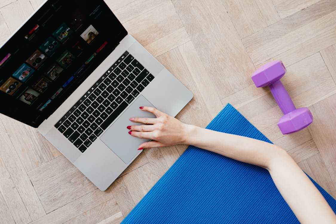 Free Top view of crop anonymous female looking for video workout courses on laptop while sitting on blue yoga mat with purple dumbbell beside on parquet floor Stock Photo