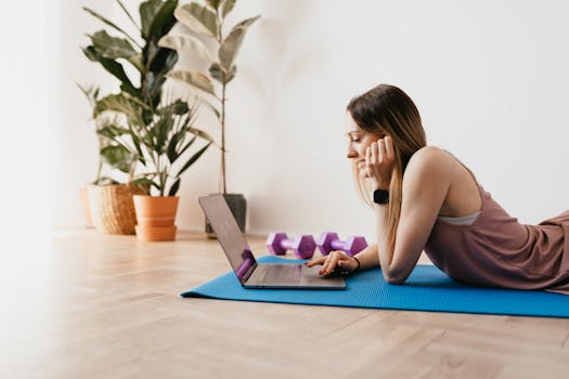 Young woman browsing on her laptop while exercising at home on a yoga mat with dumbbells nearby.