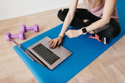 Crop female in sportswear sitting on blue yoga mat on floor and surfing internet on laptop