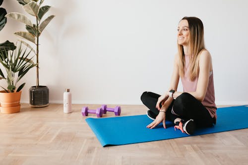 Free Full body of happy young female in activewear smiling and sitting on blue yoga mat on carpet Stock Photo