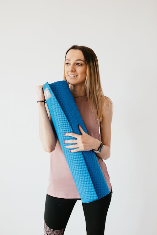 Content young female in activewear with yoga mat in hands standing near gray wall and looking away