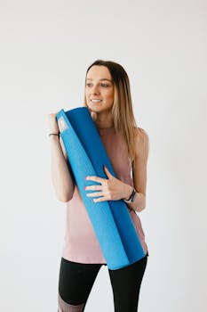 Content young female in activewear with yoga mat in hands standing near gray wall and looking away