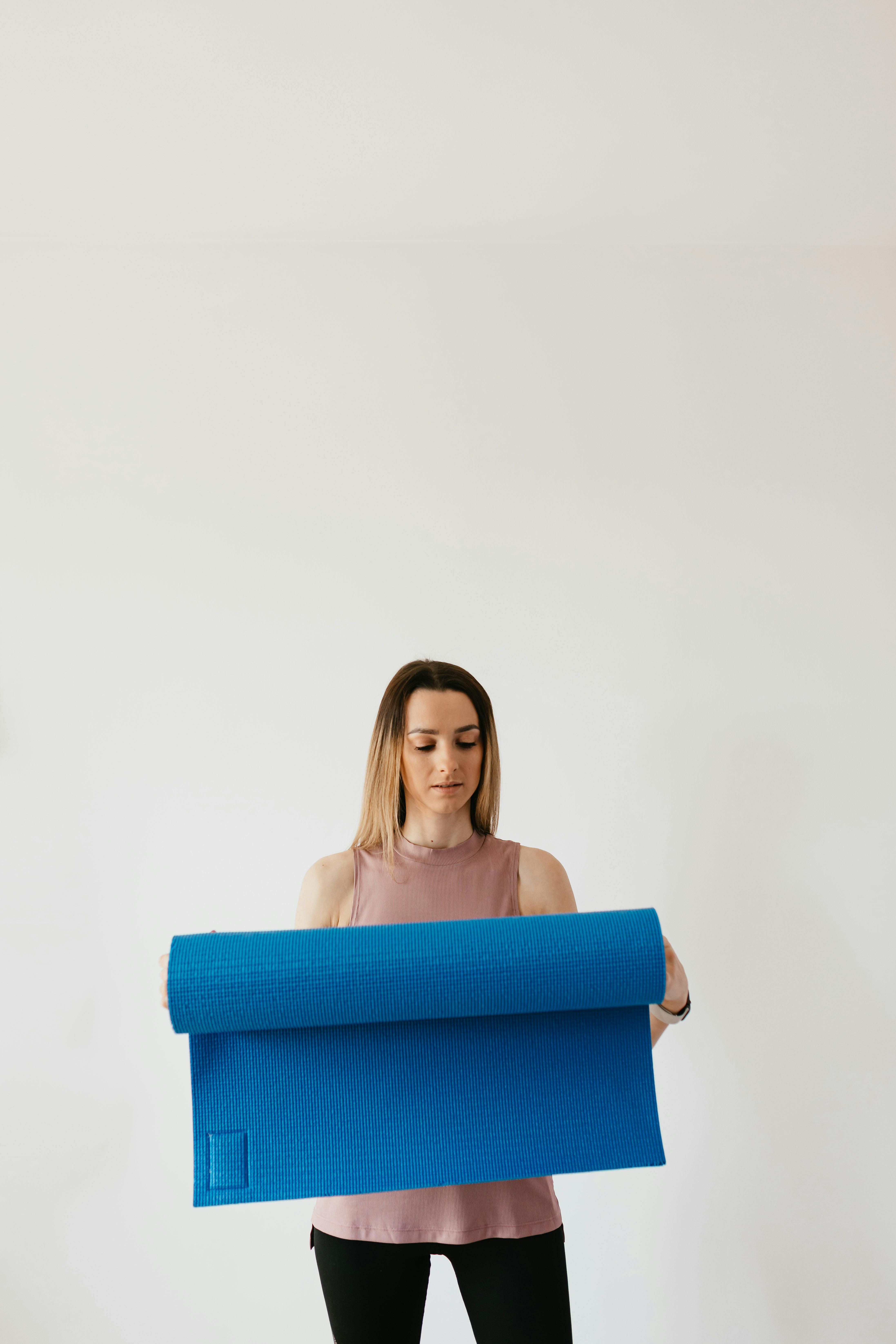 Pensive young female in activewear standing near gray wall and holding blue yoga mat in hands