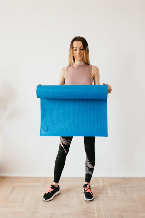 Full body young thoughtful female in activewear standing near gray wall with blue yoga mat in hands