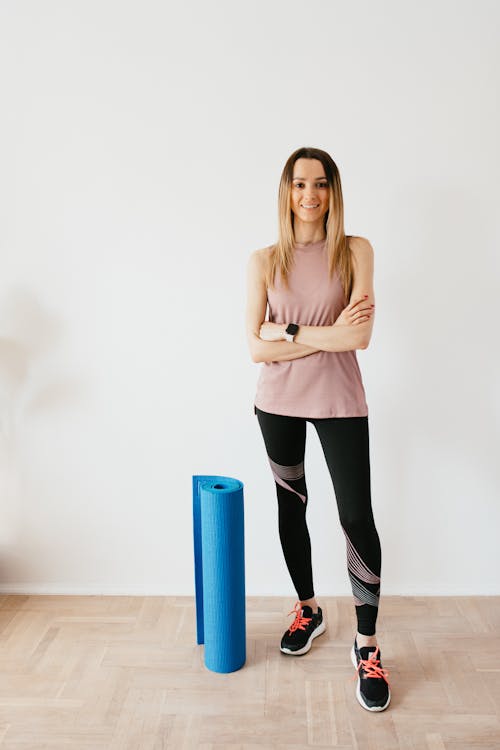 Free Full body female athlete wearing sportswear and sneakers standing against white wall with crossed arms near blue sport mat before yoga practice and looking at camera with smile Stock Photo
