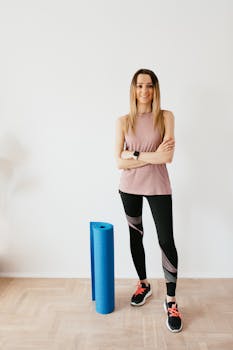 Full body female athlete wearing sportswear and sneakers standing against white wall with crossed arms near blue sport mat before yoga practice and looking at camera with smile