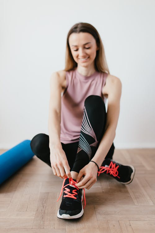 Full body satisfied young female athlete in activewear sitting on floor near folded fitness mat and tying sneakers before training while looking down with smile