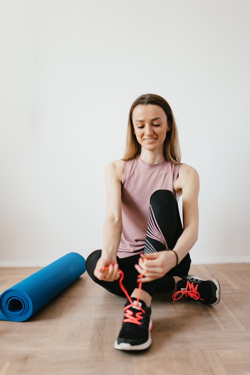 Full body positive female athlete in sportswear sitting on floor near folded blue fitness mat and tying sneakers before exercising indoors while looking down with smile