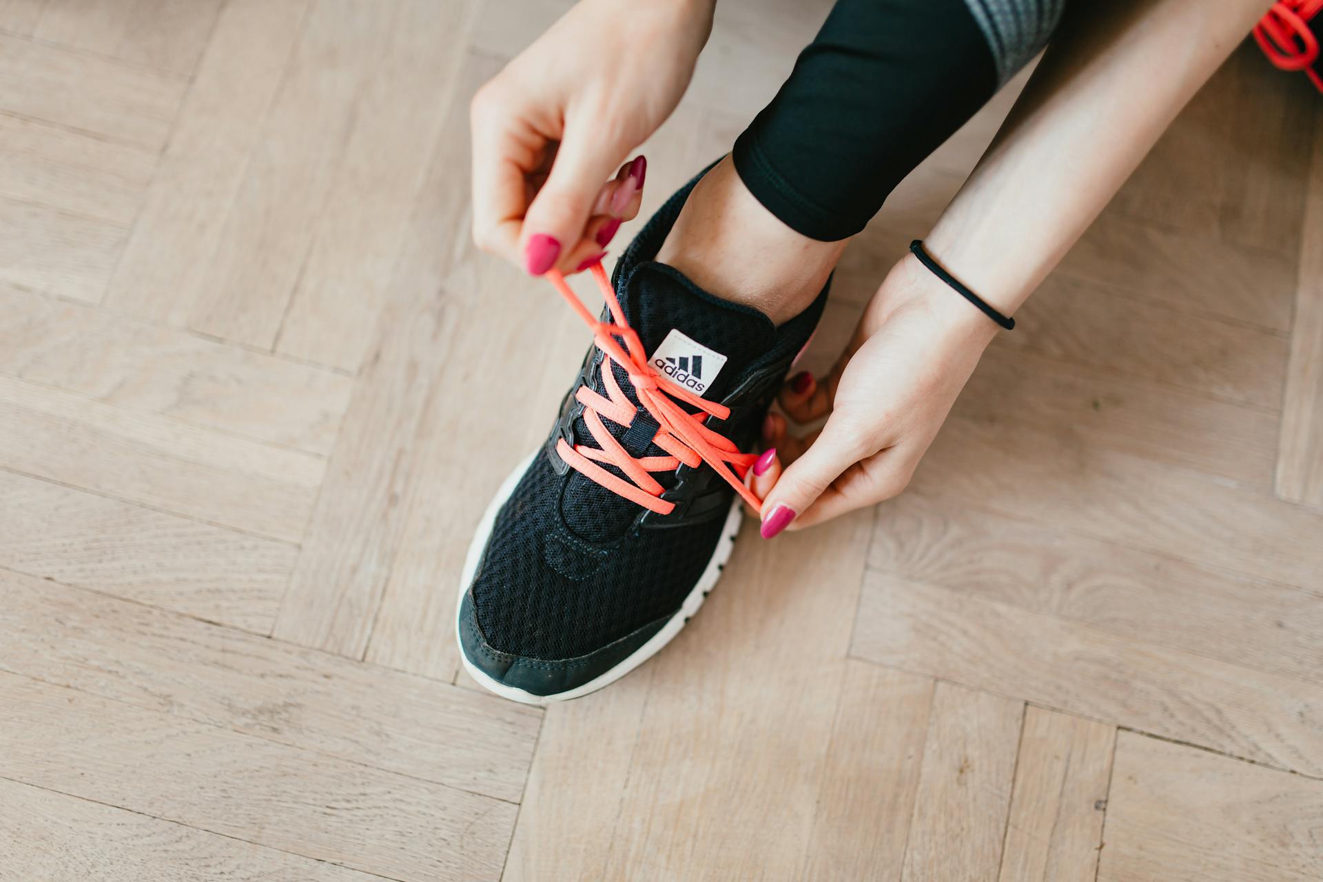 Top view of young anonymous female athlete in leggings tying shoelaces on sneakers while sitting on wooden floor in modern fitness studio and preparing for training