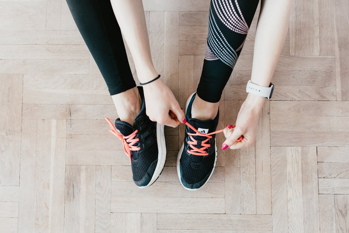 Top view of unrecognizable female athlete in black leggings tying shoelaces while sitting on wooden floor at home and preparing for workout