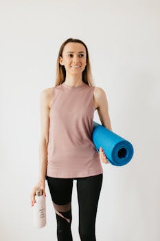 Positive young female athlete in sportswear standing isolated against white wall with blue yoga mat and water bottle while looking away with smile