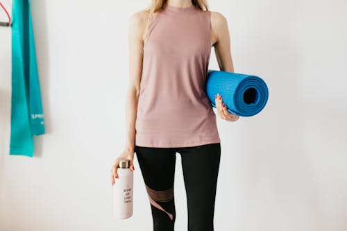 Free Faceless slim female athlete in sportswear standing with blue fitness mat and water bottle while preparing for indoors workout Stock Photo