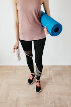 Crop fit female athlete in sportswear standing with blue fitness mat and water bottle while preparing for home exercising in morning