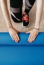 From above of anonymous female athlete in black leggings and sneakers unfolding blue mat for exercising on wooden floor in modern fitness studio