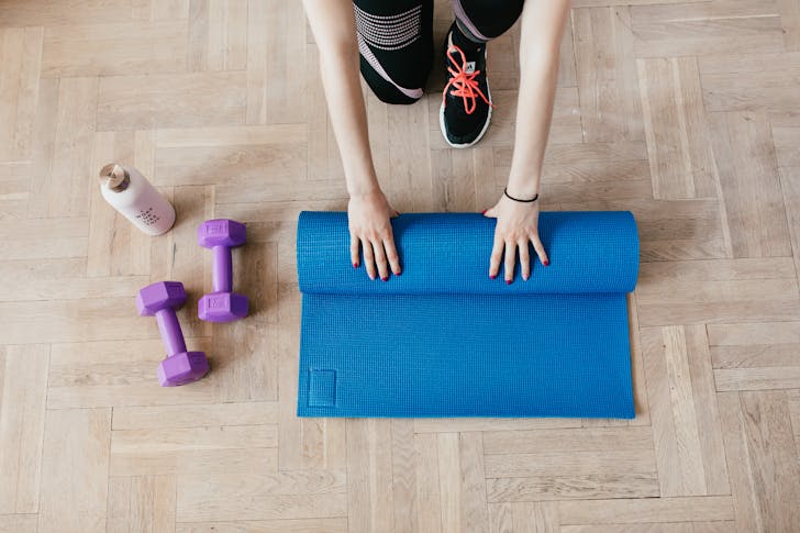 Top view of anonymous female athlete in black leggings and sneakers unfolding blue mat for exercising on floor near dumbbells and water bottle in modern fitness center