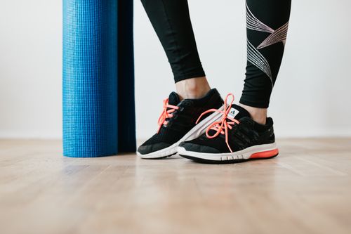 Free Unrecognizable sportswoman wearing sneakers and black leggings standing with blue fitness mat on wooden floor before exercising indoors Stock Photo