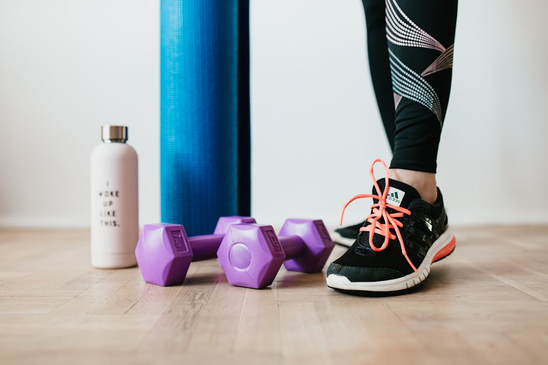 Free Crop sportswoman wearing sneakers and black leggings standing with sport mat on wooden floor near dumbbells and water bottle before exercising indoors Stock Photo