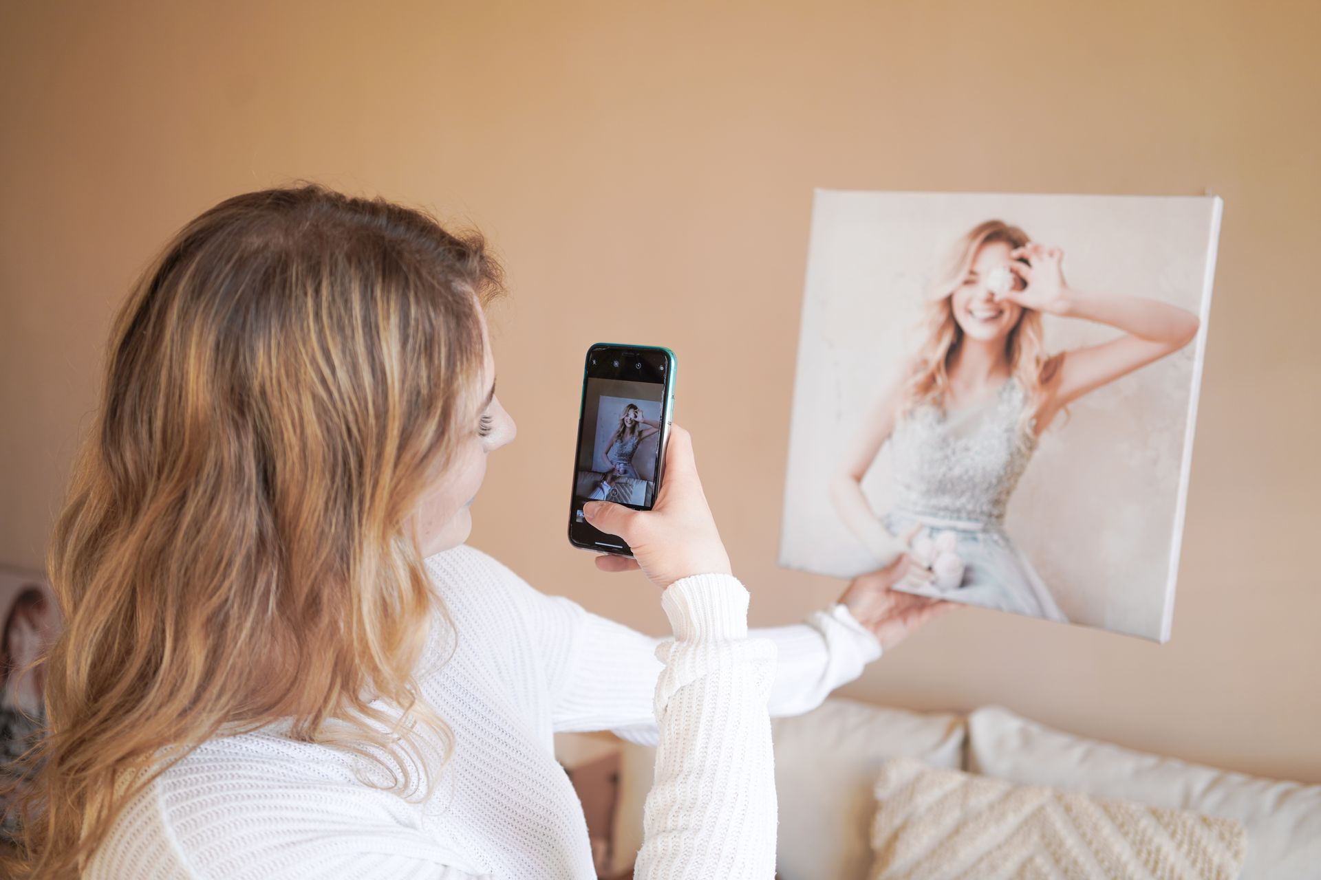 A woman captures a photo of a canvas print using her smartphone in a cozy setting.