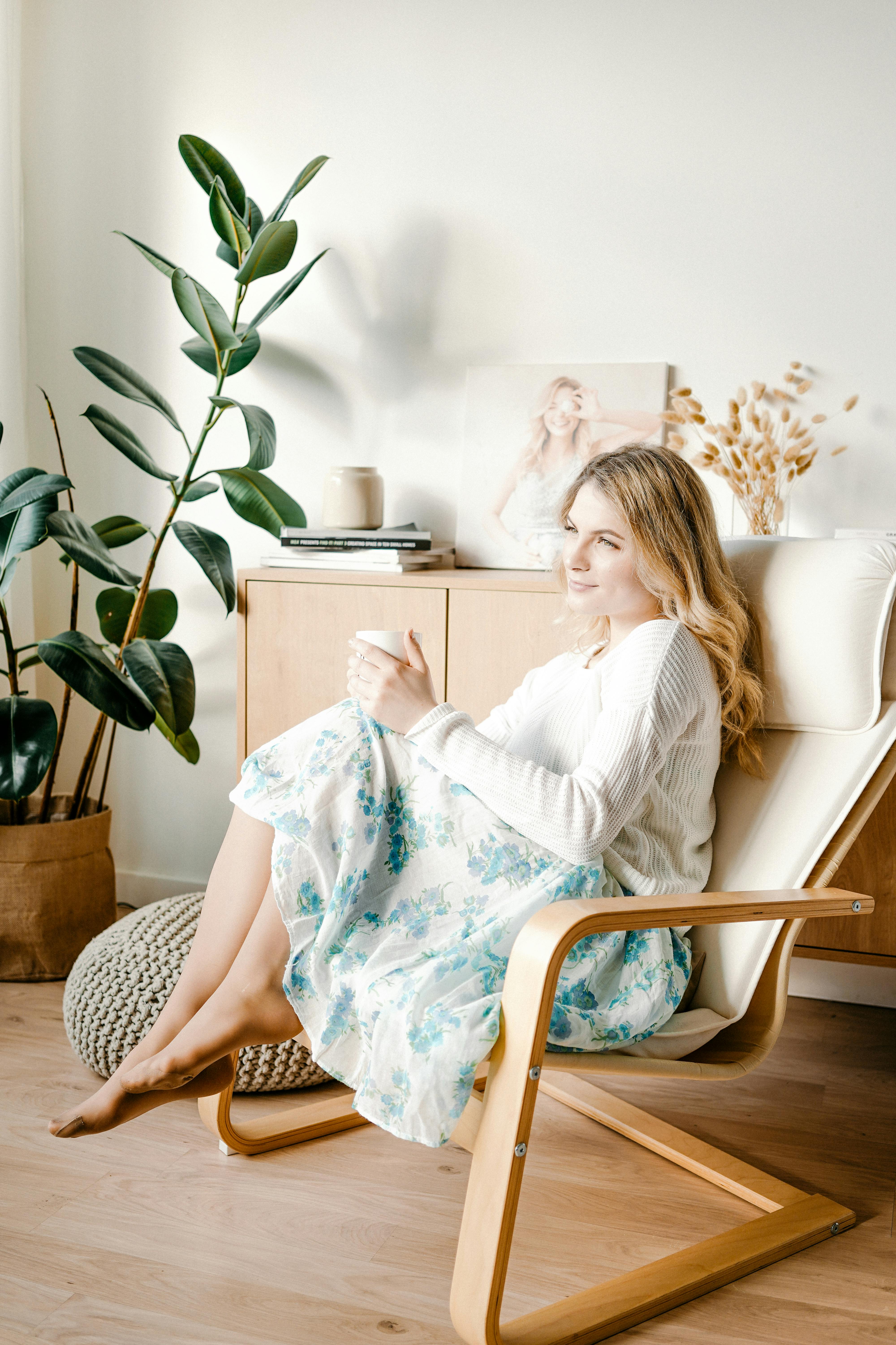 A woman in a floral skirt sits barefoot on a wooden chair, enjoying tea next to indoor plants.