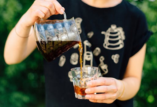 Close-up of a person pouring coffee from a pitcher into a glass outdoors.