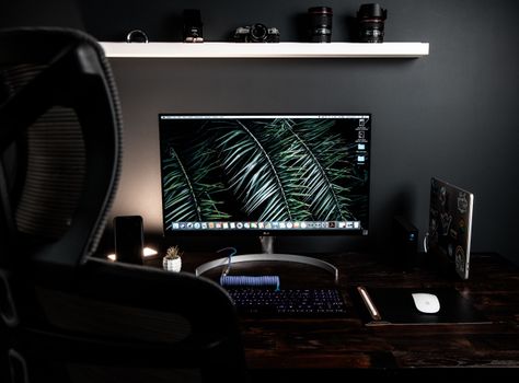 Stationery computer with keyboard and mouse on desk placed under white shelf in dark workspace with armchair near table in modern room