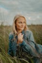 一位穿着牛仔夹克的金发女子坐在草地上，脸上带着沉思的微笑