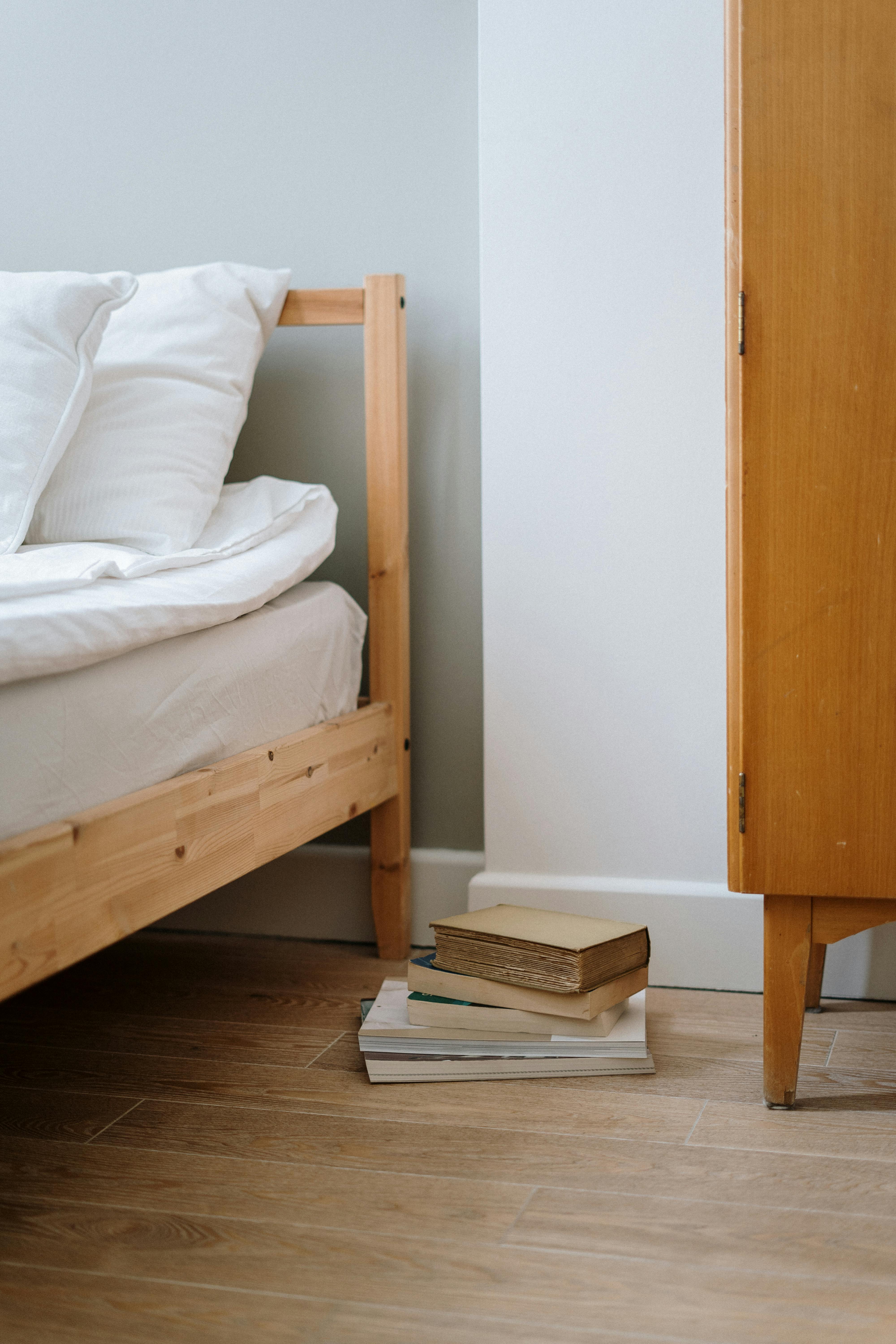 A minimalist bedroom interior featuring a wooden bed frame and a stack of books on a wooden floor, creating a cozy atmosphere.