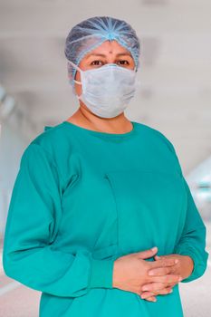 Hispanic nurse wearing scrubs, mask and hairnet, standing outdoors confidently.