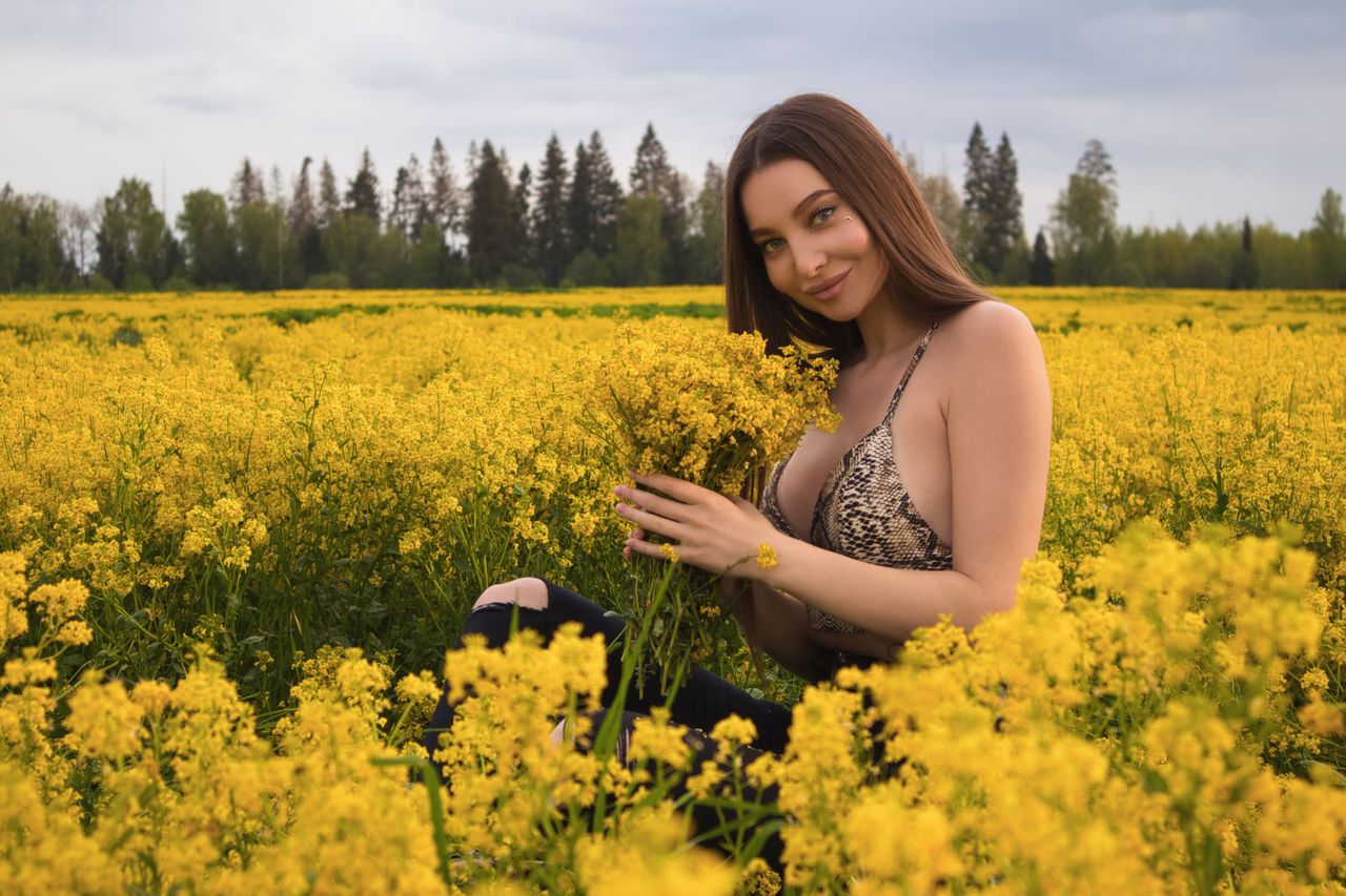 young woman sitting in a vibrant yellow flower field smiling and holdi
