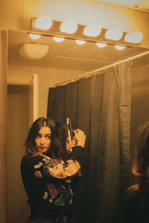 Focused female with dark hair in sweater looking in mirror and curling hair while standing near shower curtain under lamps