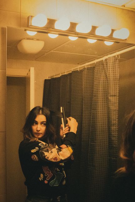 Focused female with dark hair in sweater looking in mirror and curling hair while standing near show