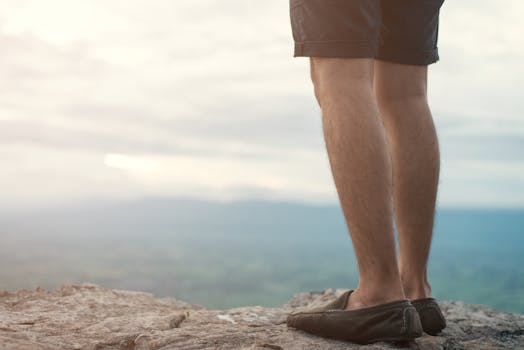 Close-up of a man's legs in casual shoes standing on a rocky edge overlooking a scenic landscape.