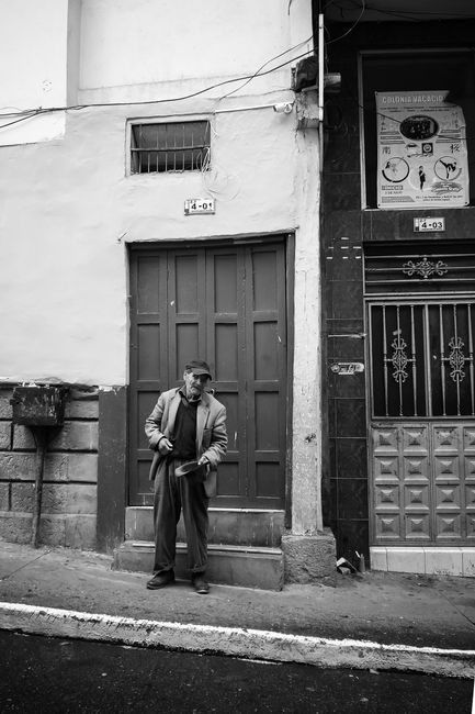 Full body of aged man in hat standing next to road on narrow pavement near shabby building with stone stairs