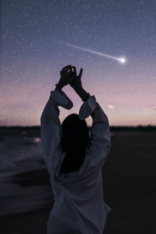 Back view of anonymous female traveler in white shirt with outstretched hands standing on sandy coast near foamy sea in twilight against starry sky