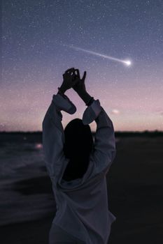 暮光之下，一位身着白衬衫的匿名女旅行者背对镜头，站在星光灿烂的大海边沙岸上，伸出双手