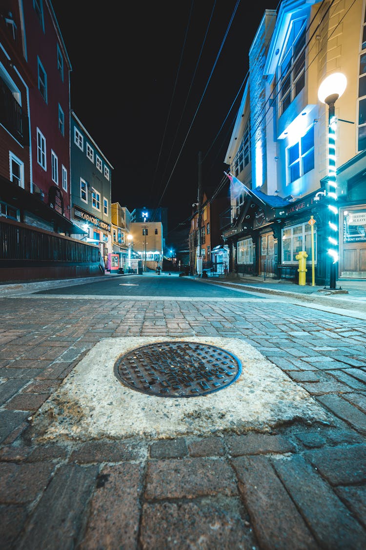 Old Manhole On Pavement Between Buildings In Town At Dusk