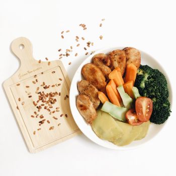 From above of plate with cooked vegetables and golden chicken slices near wooden cutting board with spilled crunchy roasted sunflower seeds