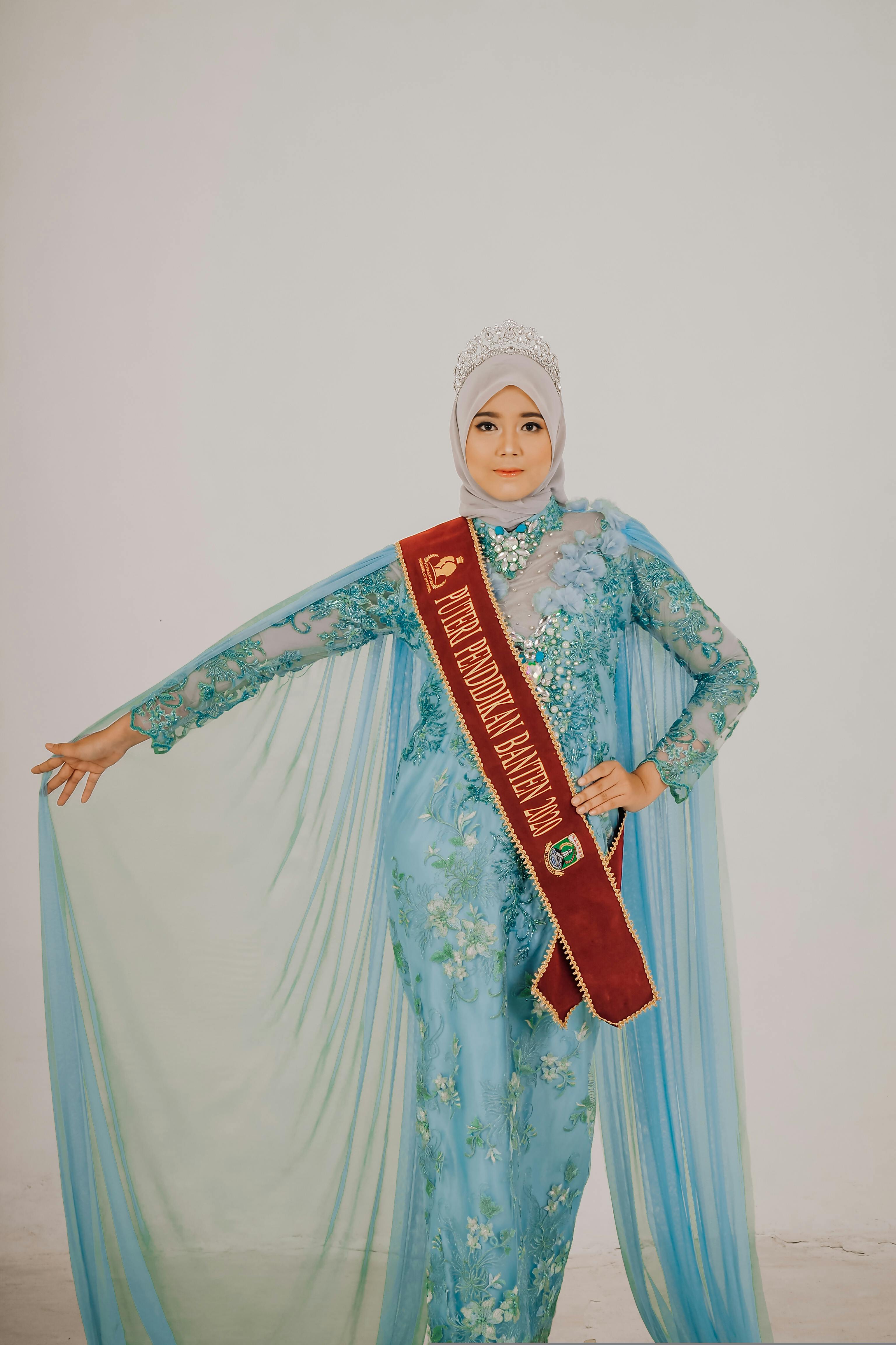 Muslim woman posing in a blue floral gown with a crown, emphasizing elegance and cultural identity.