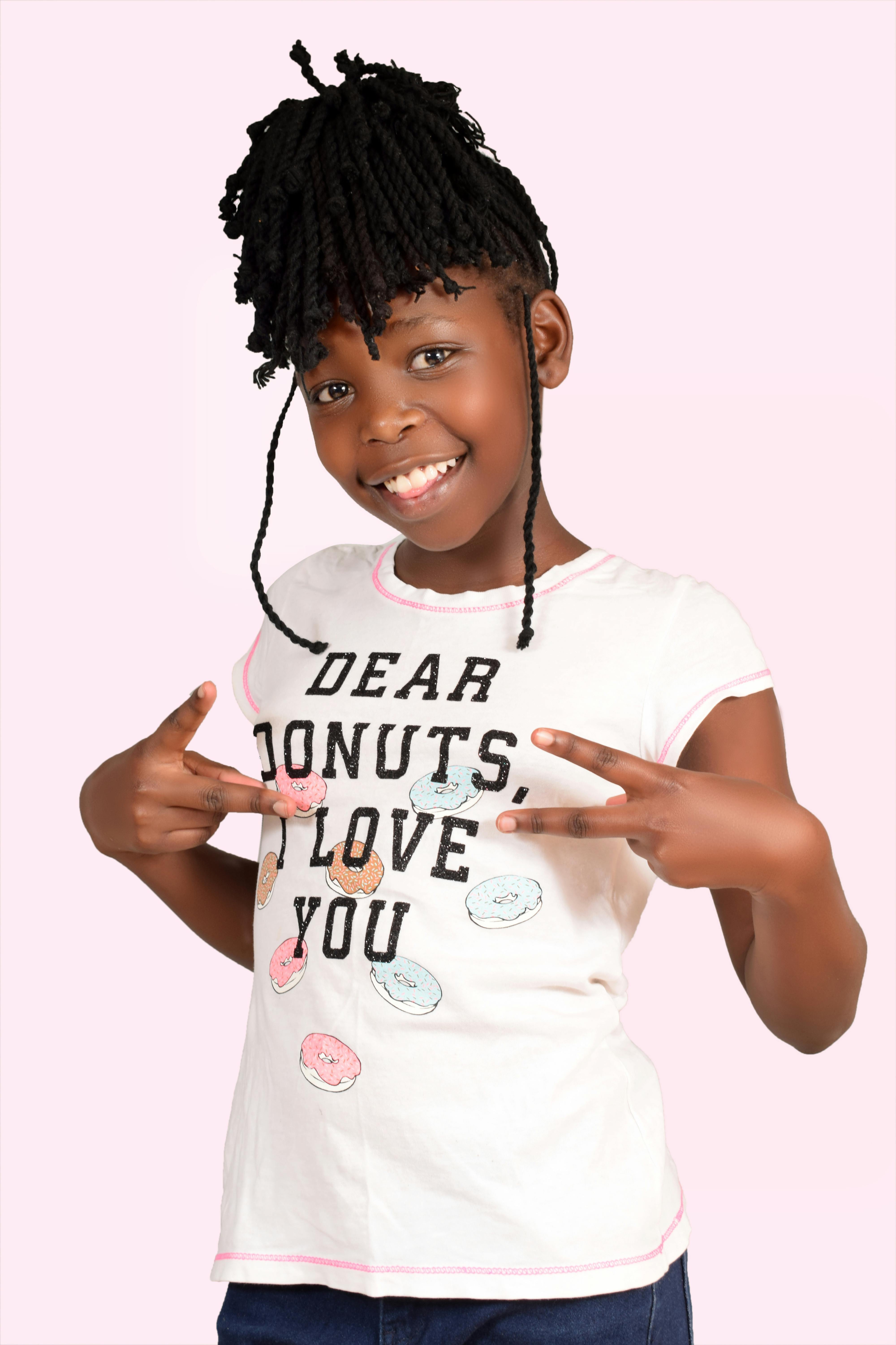 A happy child with braided hairstyle shows peace sign on a pink background.