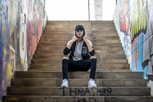 From below of young slim stylish male in hoodie and ripped jeans sitting on staircase between graffiti walls while looking at camera