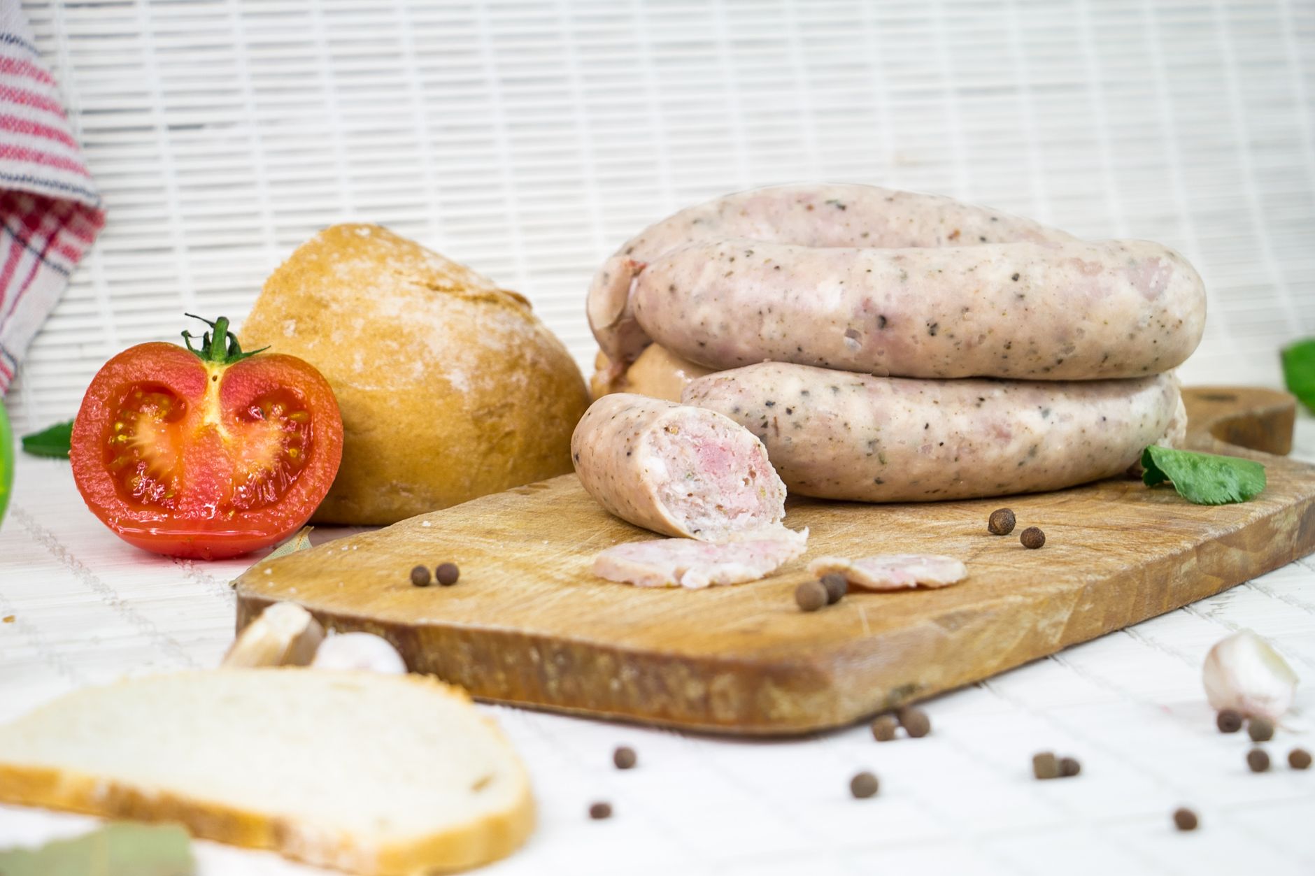 A close-up of fresh sausages, bread, and tomato on a wooden board with peppercorns.